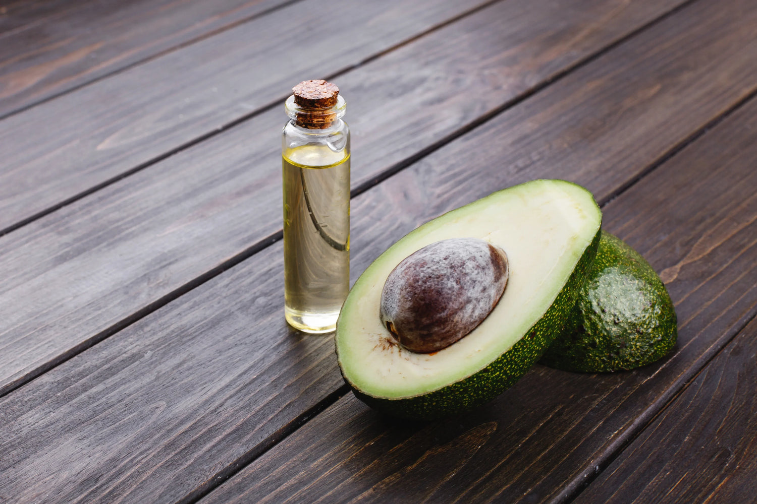 Avocado oil bottle next to a fresh cut avocado on a wooden table, illustrating a natural treatment for dry and damaged hair
