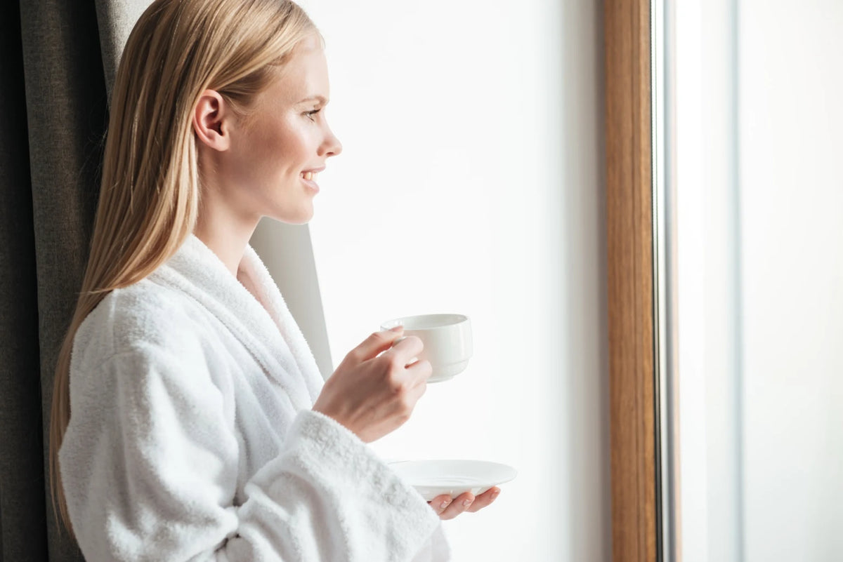 Femme souriante en peignoir savourant une boisson chaude, symbole de bien-être intérieur et d’une routine beauté naturelle avec les compléments alimentaires Biologist Mood.